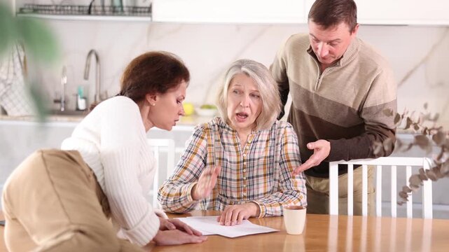 Thoughtful old woman compiling heritage document while middle-aged man and woman competing with each other in the kitchen