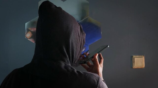 Anonymous spy person or hacker with black hoodie talking on the phone indoors. Standing near a mirror in the room. Mysterious man making phone call conversation with his back to camera. 