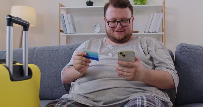 Obese man using mobile phone and credit card for payment for travel booking and tickets. Happy chubby guy sitting on living room sofa at home to pay for shopping near suitcases, using secure app