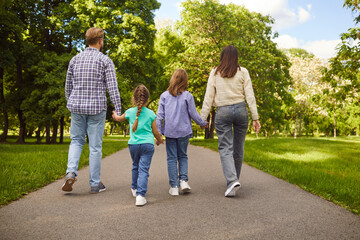 Fototapeta premium Back view of friendly family holding hands and walking along path in park, enjoying sunny weather and beautiful nature, spending time together. Parents and children being outdoors.