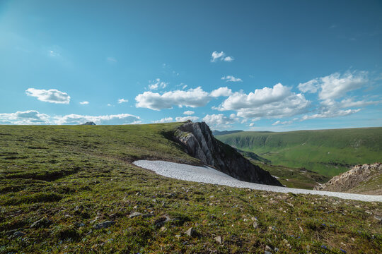 Sunlit grassy meadow, glacier and rocky cliff on precipice edge above alpine valley against big hills under clouds in blue sky in sunny day. Scenic view with green grass near sharp rocks in bright sun