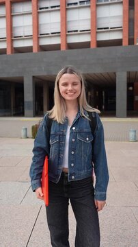 Young female student with a backpack and a red folder smiling happily. Confident woman standing in front of a modern university building