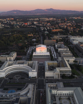 Aerial view of the Palazzo dei Congressi glows warmly against the cool tones of the surrounding buildings and distant mountains, Rome, Lazio, Italy.
