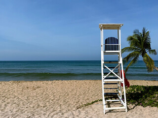White lifeguard tower on sandy beach with palm tree and blue ocean. Tropical seaside landscape with clear sky and calm sea.