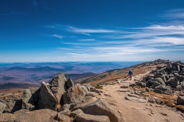 Fototapeta premium Lone hiker walking along rocky mountain trail under clear blue sky with distant rolling hills and dramatic clouds on the horizon.
