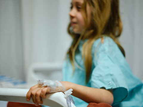 Young boy with long hair sits in a hospital bed, wearing a hospital gown and an IV drip on his hand.
