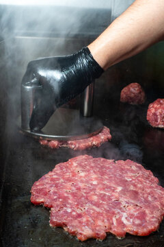 Side view of chef in black glove using heavy metal press to smash a burger patty on hot griddle