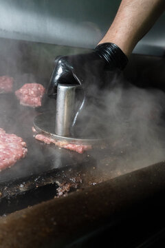 Chef's hand in black glove using metal press tool to smash a burger patty on griddle with rising steam