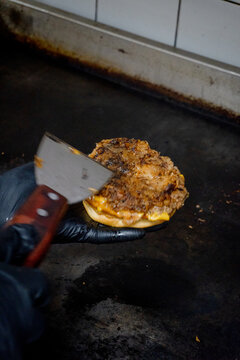 Professional chef using a metal spatula to scrape a crispy burger patty on a hot commercial griddle