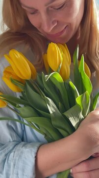 Beautiful woman enjoying the scent of fresh yellow tulips at home. Warm sunlight and soft emotions create a cozy spring atmosphere. Concept of self care, happiness, romantic mood and lifestyle.