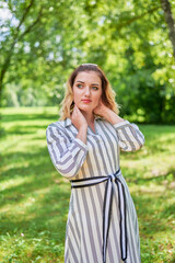 Fototapeta premium A woman in a white dress stands in a sunlit forest clearing on a summer day. In front of her is lush green grass, and behind her are tall, slender trees.