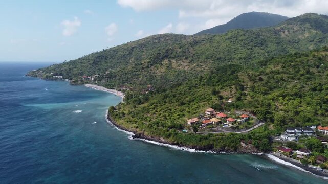 Aerial drone orbiting right transitioning from shoreline toward central green hills in Amed, Bali, Indonesia.