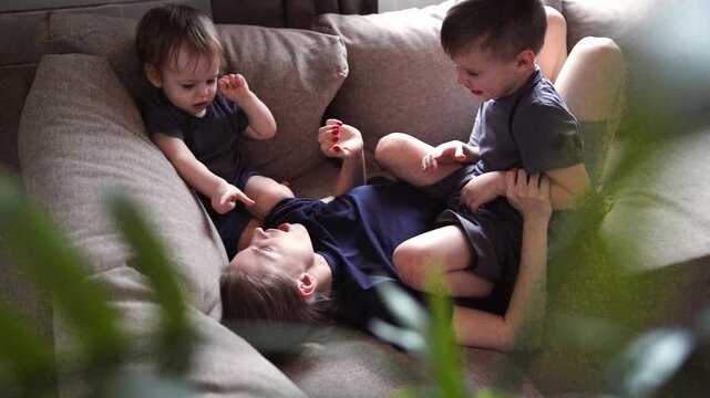 Two boys tickling white mother through plants, soft natural light, leafy foreground framing cozy sofa, candid laughter, gentle wrestling, symbolic roots and growth, nurturing family bond, peaceful