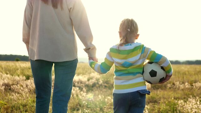 mother holding daughter hand go walk. girl walking along soccer ball. child happily smiling mother. joyful family fulfilling childhood dream walking football field sunset. represents concept family