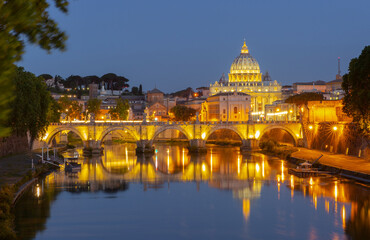 Fototapeta premium Night view of Saint Peter Basilica and bridge over the Tiber river in Rome with golden lights reflecting in calm water.
