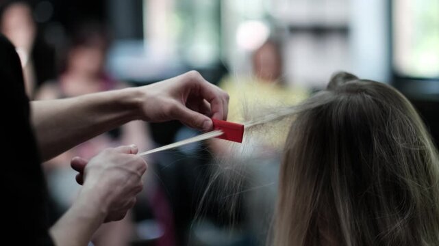 Professional hair stylist backcombing a section of blonde hair using a red comb in a busy salon