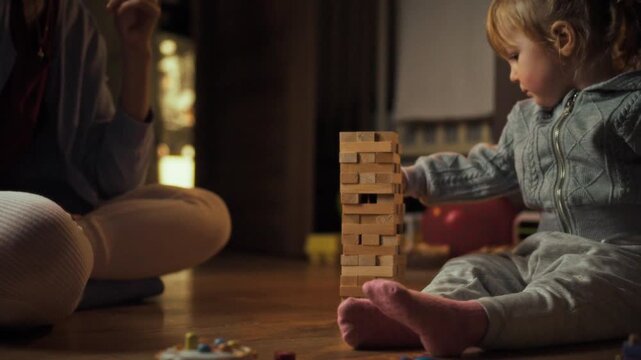 In warm evening atmosphere, mother young daughter engage playful learning floor, constructing wobbly tower from wooden Jenga blocks, promoting balance entertainment through interactive board game fun