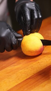 cocinero profesional con guantes de l&aacute;tex negro trocea y corta un melocot&oacute;n maduro sobre una tabla de madera, realizando movimientos precisos con el cuchillo para preparar la fruta.