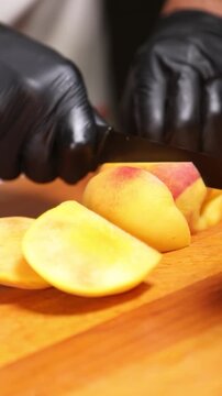 cocinero profesional con guantes de l&aacute;tex negro trocea y corta un melocot&oacute;n maduro sobre una tabla de madera, realizando movimientos precisos con el cuchillo para preparar la fruta.
