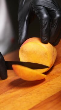 cocinero profesional con guantes de l&aacute;tex negro trocea y corta un melocot&oacute;n maduro sobre una tabla de madera, realizando movimientos precisos con el cuchillo para preparar la fruta.