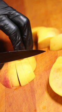 cocinero profesional con guantes de l&aacute;tex negro trocea y corta un melocot&oacute;n maduro sobre una tabla de madera, realizando movimientos precisos con el cuchillo para preparar la fruta.