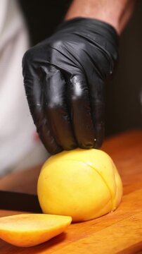 cocinero profesional con guantes de l&aacute;tex negro trocea y corta un melocot&oacute;n maduro sobre una tabla de madera, realizando movimientos precisos con el cuchillo para preparar la fruta.