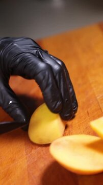 cocinero profesional con guantes de l&aacute;tex negro trocea y corta un melocot&oacute;n maduro sobre una tabla de madera, realizando movimientos precisos con el cuchillo para preparar la fruta.