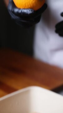 Camara lenta de cocinero profesional con guantes de l&aacute;tex negro toma una naranja fresca de un frutero, seleccion&aacute;ndola como ingrediente para el siguiente paso de la preparaci&oacute;n.