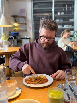 Man enjoying potato pancakes and mushroom sauce at wooden cafe table. Everyday restaurant dining, urban lifestyle routine, comfort food culture and relaxed social atmosphere.