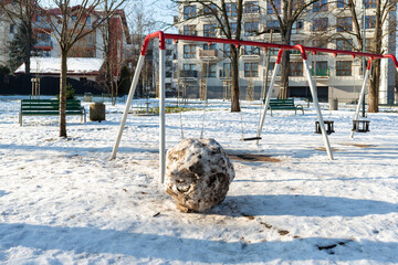 Metal swing set in a snowy park with a large dirty snowball standing in front. Bare trees and apartment buildings frame the winter playground scene showing seasonal outdoor environment during cold wea © Volha