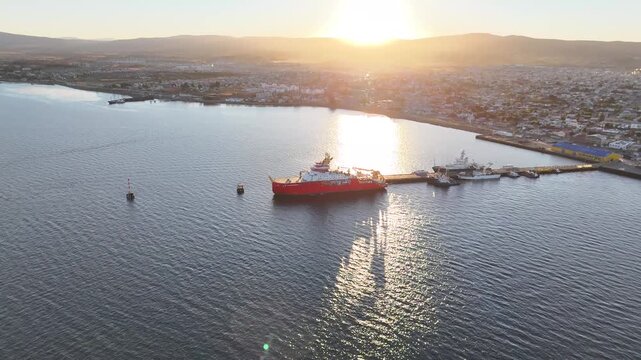 Aerial moves toward the red polar research ship RRS Sir David Attenborough at sunset in Punta Arenas, tilting down for a detailed top-down view of the deck and helipad