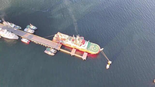 Aerial rises and pulls back from the RRS Sir David Attenborough at Prat Pier, revealing the coastal promenade, city waterfront, and ships anchored in the Strait of Magellan