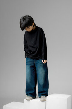 Asian boy wearing black long sleeve shirt and blue denim jeans standing on white studio blocks looking down, kids fashion portrait.