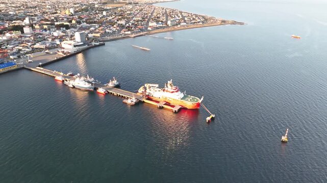 Aerial circling the red polar research ship RRS Sir David Attenborough at sunset, showing the vessel docked at Prat Pier with the Punta Arenas coastline in the background