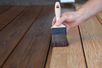 Fototapeta premium Brown oil for protection of wood by staining. Painting a larch for protecting and pests. Woodworker applying finish to terrace deck board. Repairman varnishing with brush wooden plank floor surface.