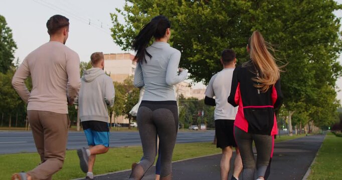 Marathon run of runners jogging together on road in summer park, back view. People practice sport exercises outdoors for wellbeing, young athletes in sportswear running with unity and motivation