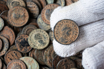 Gloved hand holding an ancient Roman bronze coin. Close up of a numismatic artifact with profile of emperor Valentinianus. Pile of old coins in background. Archaeology concept © scaliger