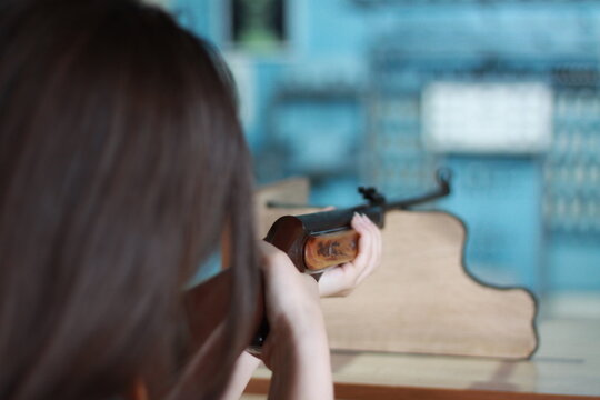 A back-view, close-up shot of a dark-haired girl holding a classic wood-stock air rifle. She is focused on aiming at a target in a soft-focus indoor shooting gallery. The image captures themes of prec