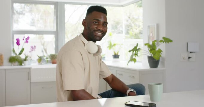 Man sitting at kitchen counter with headphones and mug, camera recording, straightening and smiling