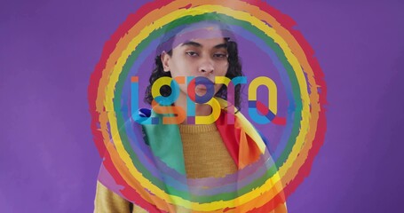 Posing person wearing mustard sweater draping pride flag on purple wall with ring overlay letters