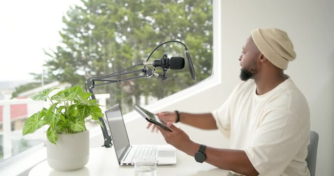 Man wearing beanie recording from tablet by window at round table with microphone, copy space