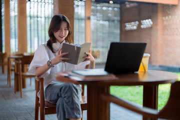 Fototapeta premium Asian young woman reading book peacefully in modern cafe environment headphones around neck laptop nearby for work or study