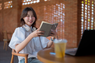 Fototapeta premium Asian young adult woman reading book enjoying leisure time learning studying cafe workspace relaxed smiling girl headphones laptop drink modern brick wall background