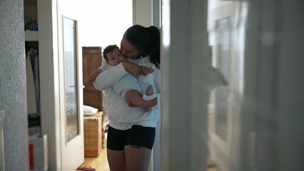 Fototapeta premium Young mother holding her baby in a warmly lit home hallway, cradling the infant gently. She kisses the baby’s head while standing near an open door