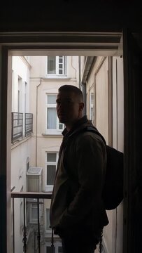 Vertical video: Pausing man turning toward courtyard at apartment doorway, settling backpack straps