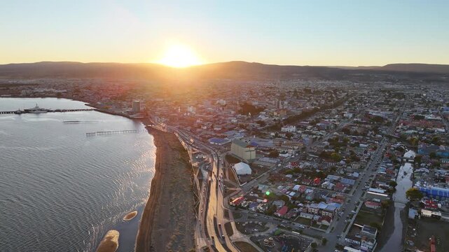 Aerial camera pans left over the coastal road and colorful rooftops of Punta Arenas, Chile, as the sun sets behind the Patagonian hills, reflecting on the Strait of Magellan