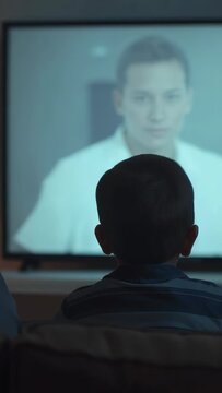 Vertical video: Watching child in striped shirt sitting on couch TV showing blurred man in white