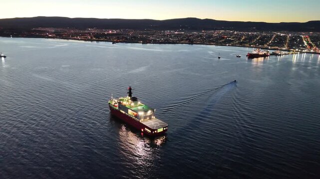 Aerial pulls out from the illuminated RRS Sir David Attenborough research vessel, revealing its wake in the Strait of Magellan and the twinkling city lights of Punta Arenas, Chile