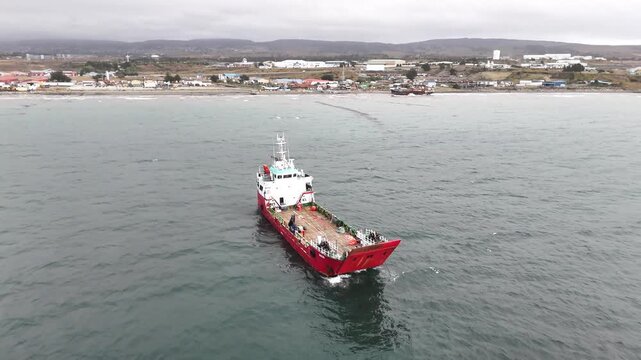 Aerial pulls back from a close-up of the barge Anan, moving out to reveal the vessel in the Strait of Magellan with the industrial coastline of Punta Arenas in the background