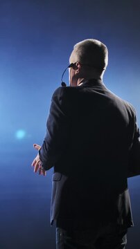 Senior male motivational speaker passionately gestures while delivering an inspiring speech on stage, wearing a headset microphone. Vibrant stage lights illuminate the scene, creating a dynamic and